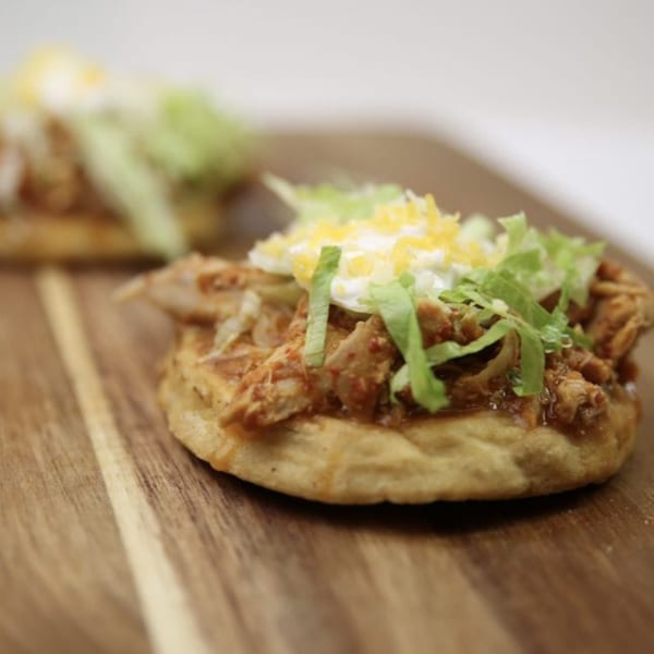 Two Mexican sopes with shredded chicken, lettuce, cheese, and sour cream on a wooden board.