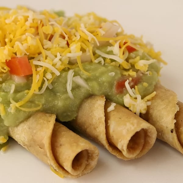 Three crispy taquitos topped with guacamole, shredded cheddar, and white cheese.