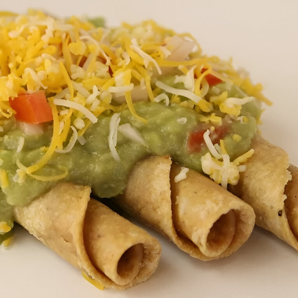Three crispy taquitos topped with guacamole, shredded cheddar, and white cheese.